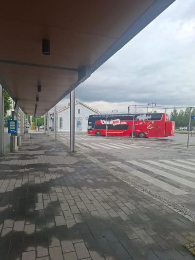 The image depicts a bus stop in Mikkeli, Finland, under a cloudy sky. A red double-decker bus with "Onnibuss.com" branding is parked on the road, partially obscuring a pedestrian crossing. The bus stop itself features a covered waiting area with multiple pillars and overhead lighting. The pavement is made of dark grey rectangular pavers, some of which are wet, suggesting recent rain. In the background, a light blue building with white trim and several windows is visible, along with some trees. A blue ticket vending machine is mounted on a pole to the left of the bus stop. The overall scene is quiet, with no people visible.