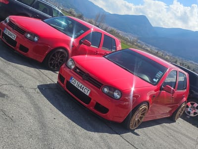 The image depicts two red Volkswagen Golf IV cars parked side-by-side in Kraynitsi, Bulgaria. The front car has a Bulgarian license plate "CO 1644 CT". The vehicles are likely on an asphalt surface, possibly a parking lot or road shoulder. The background includes a distant view of mountains and a valley. The sky is partly cloudy.