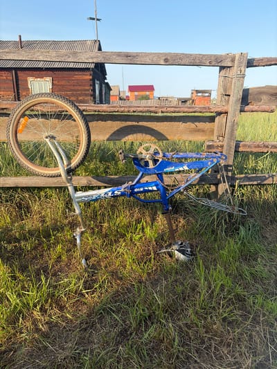 The image depicts a derelict blue bicycle leaning against a weathered wooden fence in a rural setting. The bicycle is missing its rear wheel and has its front wheel and handlebars propped on a wooden beam of the fence. The frame of the bicycle is blue with white lettering that appears to read "STELS 510". The front wheel is a spoked bicycle wheel with a black tire and an orange reflector. The bicycle is positioned in overgrown green grass.
In the background, a rustic wooden house with a dark, corrugated roof is visible. The house has a small window with white decorative trim. Further in the background, there are other buildings with red roofs and what appears to be a rudimentary wooden structure. The sky is clear and blue, indicating a sunny day.
The scene appears to be in or near the village of Myandigi, Russia, given the architectural style of the buildings and the natural environment. There are no people or animals visible in the image. The overall impression is one of abandonment and rural decay.