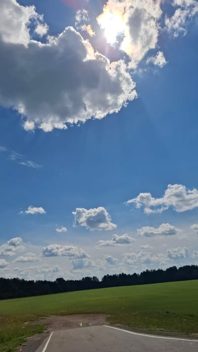 A bright, clear blue sky dominates the upper portion of the image, with a few fluffy white clouds scattered across it. The sun is prominently visible, partially obscured by a large, dense cloud formation on the left side, casting rays of light and some orange and pink hues onto the surrounding clouds. The lower half of the image shows a vibrant green field, stretching towards a dark line of trees in the distance. In the foreground, a section of a paved road or path is visible, with the edge of it cut off by the frame. The scene appears to be a rural or semi-rural outdoor setting, likely in Stantsionno-Oyashinskii, Russia, given the context provided. There are no people or animals visible in the image.