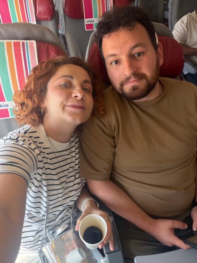 A couple is posing for a selfie on what appears to be an airplane. The man has dark curly hair, a beard, and is wearing a brown t-shirt. The woman has reddish-orange curly hair, is wearing a black and white striped shirt, and is holding a cup of coffee and a silver handbag. There are airplane seats and airline branding visible in the background. The location is possibly a flight from or to Tzafria, Israel, but there is no confirmation in the image.