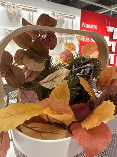 The image shows a close-up view of an autumnal arrangement of artificial leaves and pine branches inside a white ceramic pot, which is placed within a wire basket. The leaves are in various shades of red, orange, yellow, and brown, with some appearing to be maple or oak leaves, and others more rounded like eucalyptus. A small, dark, pinecone-like object is nestled among the greenery, seemingly held together with a white adhesive. The background is blurred but suggests an indoor retail environment. A bright red sign with white text reading "Nuestro" is visible, partially obscured by the arrangement. This sign, and the retail context, implies a commercial setting. Based on the information provided, this image was captured in the city of Siero, Spain, likely in a store or showroom.