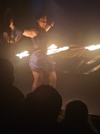 A woman in a dark, sparkling dress is performing with fire. She is holding two flaming objects, spinning them around her body. The background is dark, suggesting a nighttime or indoor setting. The bottom of the frame shows the silhouettes of audience members, indicating this is a performance viewed by others. The lighting is focused on the performer and the flames, creating a dramatic effect.