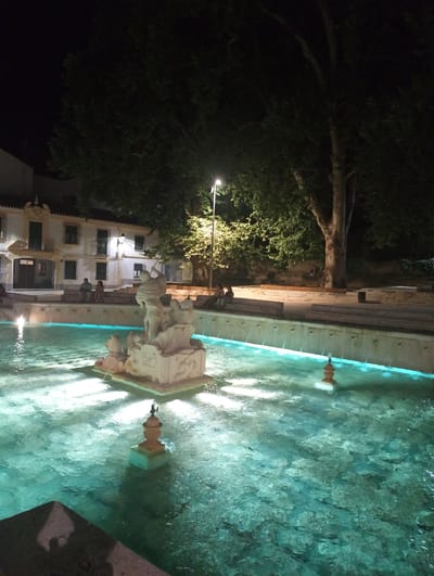The image captures an outdoor fountain scene at night, likely in Priego de Córdoba, Spain. The fountain's water is illuminated with a turquoise glow from submerged lights, creating a reflective surface. A white stone sculpture depicting figures, possibly mythological or historical, stands in the center of the fountain. Adjacent to the fountain, a large tree with dense foliage dominates the background. Behind the fountain, a white building with several windows and balconies is visible, and a few people are seated on steps leading up to it. A street lamp illuminates a portion of the area, casting light on the building and the tree. The overall atmosphere is tranquil and serene.