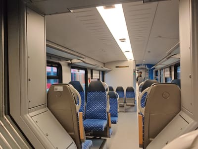 The image shows the interior of a modern train carriage. The seats are upholstered in a blue and white patterned fabric, with black headrests and light gray armrests. The floor is a light gray laminate, and the walls and ceiling are also light gray. The carriage is illuminated by long, rectangular LED light panels in the ceiling. In the background, there is a glimpse of a person wearing a dark jacket and a cap, standing near a doorway. The scene appears to be within the city of Memmingen, Germany, suggested by the train's interior and the presence of a QR code on a sticker that may relate to local transport information.