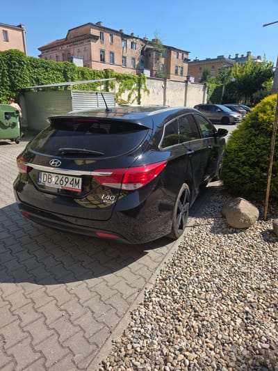 In the city of Brzeg, Poland, a black Hyundai i40 station wagon, license plate DB 2694M, is parked on a paved area adjacent to a gravel patch and a dense green bush. The car is facing away from the viewer, and its rear is visible. The license plate is clearly legible. To the left of the car, a green dumpster is situated near a corrugated metal fence partially covered in ivy, which climbs along a textured wall. In the background, two old buildings stand under a clear blue sky. One building appears to be in disrepair with missing window panes, while the other, slightly further back, is a lighter color with a more intact facade. A person, dressed in light-colored clothing, is partially visible on the left side of the image, near the dumpster. The overall scene suggests an outdoor parking area, possibly in a residential or commercial district.