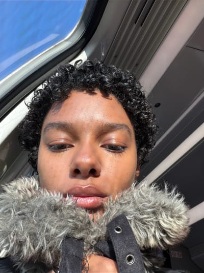 A young individual with dark skin and short, curly black hair is depicted in a close-up, upward-angled portrait, likely a selfie. The subject's eyes are lowered, and lips are full and glossy. They are wearing a dark jacket featuring a prominent, fluffy grey and brown faux fur collar, which partially covers the lower face. Two silver grommets are visible on the jacket's left side, with a hand partially obscured, appearing to hold the collar. The background indicates an interior of a public transport vehicle, likely a train or bus, in Chelles, France. A clear blue sky is visible through a window in the upper left, suggesting bright, sunny outdoor conditions. Strong sunlight illuminates the subject's face and casts distinct shadows on the light-colored, ridged overhead paneling of the vehicle's interior.