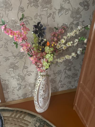 A vase of artificial flowers is displayed against a floral patterned wallpaper. The vase is tall and cream-colored with an ornate, cut-out design featuring floral motifs. The floral arrangement includes pink, yellow, white, and green artificial flowers, some with small dark berries. The wallpaper behind the flowers has a textured, light-colored floral pattern with a hint of silver. To the right of the vase, a portion of a wooden door, painted a light peach color, is visible. In the foreground, a section of a patterned rug with brown, beige, and black colors is visible on a brown floor. The image appears to be taken indoors. No people are present in the scene. The location provided, Yazykova Pyatina, Russia, is noted but cannot be verified from the visual information within the image itself.