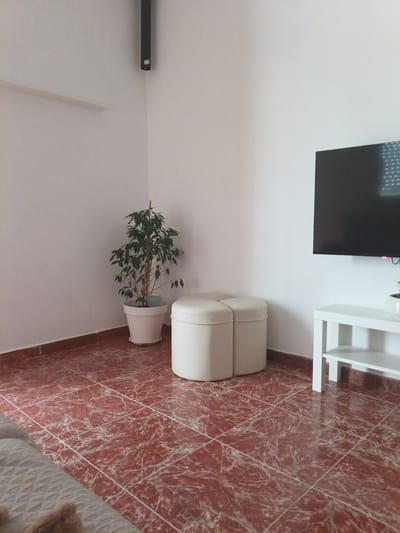 The image shows a domestic scene within a room, likely a living area. The main subjects are a large potted plant with lush green foliage, a light-colored upholstered ottoman composed of two connected sections, and a television mounted on the wall. To the right, a white media console with shelves is visible, holding a small potted plant. The floor is covered in red and white marble-patterned tiles. The walls are painted white, and a dark, cylindrical object, possibly a speaker or projector, is mounted high in the corner where two walls meet. The room appears to be well-lit, suggesting daylight, although the specific time of day is not discernible. There is no discernible activity taking place, presenting a static snapshot of the room's arrangement. The overall impression is one of a modern, minimalist interior. The location context provided is Campillo de Altobuey, Spain. No visible text is present in the image.