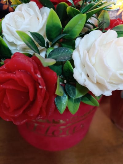 The scene, located in Asaka, Uzbekistan, depicts a close-up view of an artificial flower arrangement. The arrangement features several red and white roses, appearing to be crafted from a wax or soap-like material, intermingled with green artificial leaves. These flowers are contained within a deep red, rounded pot. The word "Flower" is visible in stylized gold lettering on the pot's side. The pot rests on a light brown surface, consistent with wood grain. A blurred, dark reddish object is partially visible in the background to the right. No individuals or discernible actions are present in the image, which suggests an indoor environment.