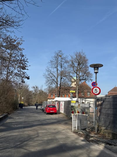 The scene depicts a daytime outdoor setting in Bad Nauheim, Germany, under a clear blue sky with two faint contrails. An asphalt path extends into the background, bordered on the left by leafless deciduous trees and bare brush, and on the right by a temporary metal construction fence. A lone individual, dressed in dark clothing, walks away from the viewer on the asphalt path.
On the right side of the path, a red compact car with the license plate "FB SS 100" is parked facing away from the camera, adjacent to the construction fence. Other parked vehicles are partially visible further along the fence line. A white, hinged barrier gate marks an entry point to the fenced area. A dark-colored street lamp with a circular, flat top stands upright near the fence, accompanied by two circular traffic signs: a red-bordered "5" speed limit sign and a "No Stopping" sign. A black rectangular sign with "VIDEO GUARD" in yellow text is mounted on a pole behind the fence. The ground bordering the fence line consists of disturbed earth and gravel, with a metal grate visible.
On the left side of the path, a large, dark blue or black waste container with yellow elements is positioned further down. Numerous bare deciduous trees are visible throughout the image, with a prominent cluster of mistletoe located in one of the taller trees behind the construction fence. In the background, beyond the trees, several buildings with traditional orange-tiled pitched roofs are discernible, including one central structure featuring a light-colored tower with a dome-like top. Direct sunlight casts discernible shadows on the asphalt path.
