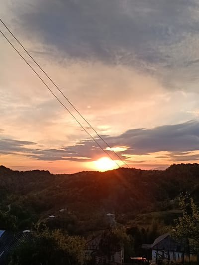 The image depicts a sunset over a hilly landscape in Yuada-Gulrypsh, Abkhazia. The sky is filled with a mix of orange, pink, and gray clouds. The sun is setting behind the hills, casting a warm glow over the scene. Two power lines stretch across the upper part of the image. In the foreground, there are some buildings and trees. The overall impression is one of a peaceful, rural setting.