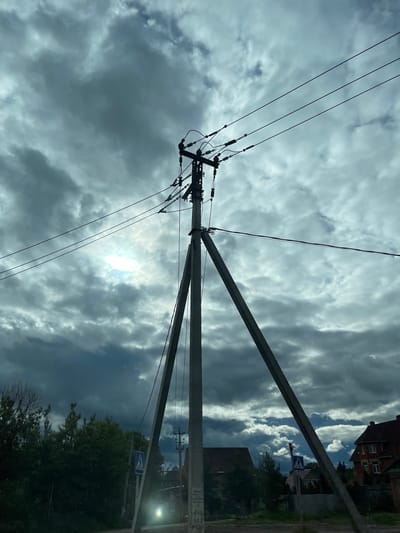 The image shows a utility pole with power lines extending upwards into a cloudy sky. A crosswalk sign is visible along with some buildings, trees, and street lights. Based on this evidence, the image appears to be in an urban or suburban area, possibly Semyonovskoye, Russia, as specified. No people are visible. The focus is on the infrastructure and sky.