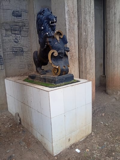 The scene depicts a weathered, dark-colored lion statue located outdoors at Itakpe Junction, Nigeria. The statue, appearing to be made of a dense material, stands erect on its hind legs, with its front paws resting on a dark, shield-like object accented with gold-painted scrolls. The lion's mouth is open, revealing molded teeth, and its black surface shows signs of paint degradation and lighter wear marks, particularly on the mane and raised details. It rests on a low, rectangular pedestal, approximately 0.7 meters high, clad in off-white square ceramic tiles exhibiting significant dirt, grime, and brownish discoloration, especially near the base. The immediate ground surrounding the pedestal is uneven, dry, reddish-brown earth mixed with small stones, fragmented debris, and scattered litter, including a discarded white wrapper and a small orange packet. In the background to the left, a light-colored wall surface is extensively covered with black marker graffiti, depicting various grid-like patterns and illegible scribbles. To the right, multiple large, vertical, fluted columns, appearing to be concrete or stone, rise upwards, showing weathering and discoloration. No individuals or active interactions are observed in the image.