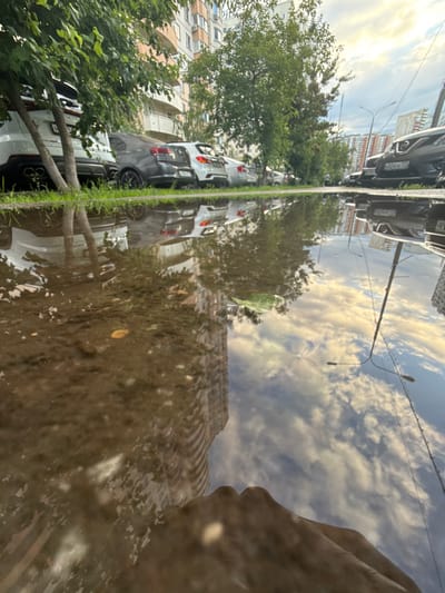 The image shows a street scene in Putilkovo, Russia, with a large puddle in the foreground reflecting the sky and surrounding buildings and trees. Several cars are parked along the street, including white, gray, and black vehicles. A tall apartment building, some trees with green foliage, and streetlights are visible. The sky is partly cloudy with white clouds against a blue sky, illuminated by sunlight. The reflection in the puddle is distorted but shows the inverted images of the sky, buildings, trees, and streetlights. In the bottom right corner of the puddle's reflection, a portion of a person's hand is visible, with fingers extended. The ground around the puddle appears to be asphalt or pavement, with some grass on the left side of the frame.