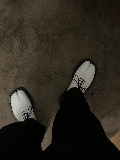 From a high angle, white, patent leather tabi shoes with black laces and contrasting soles stand out against a textured, grey carpet. Dark, wide-legged trousers obscure parts of the shoes, creating a stark contrast in color and texture. The composition focuses on the distinct footwear.
