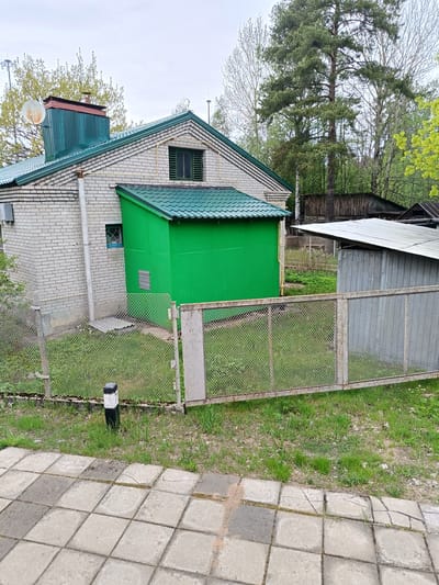 The image shows the exterior of a brick building with a green extension in Ushkovo, Russia. A fence runs along the perimeter, separating the building from a paved area in the foreground. The building features a green roof and a chimney with a satellite dish attached. Trees are visible in the background, and a smaller structure is situated to the right of the main building within the fenced area. The overall scene suggests a residential or rural setting.