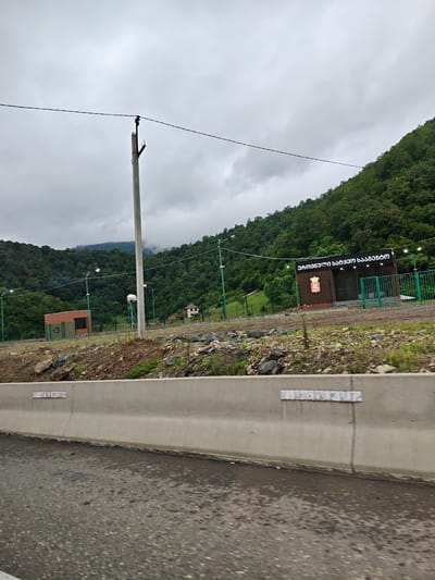 The image captures a roadside scene in Grigalati, Georgia, characterized by a grey, overcast sky and lush, green mountains in the background. A concrete barrier runs horizontally across the foreground, separating the paved road from the raised embankment. On the embankment, a concrete utility pole stands prominently, with a power line extending across the frame. Beyond the pole, a cluster of buildings and fencing is visible. One structure appears to be a small, reddish-brown building with a green fence, while another is a darker structure with a sign featuring Georgian script and a coat of arms. The surrounding terrain is rocky and covered with sparse vegetation. The overall atmosphere is subdued, typical of a cloudy day in a mountainous region.
