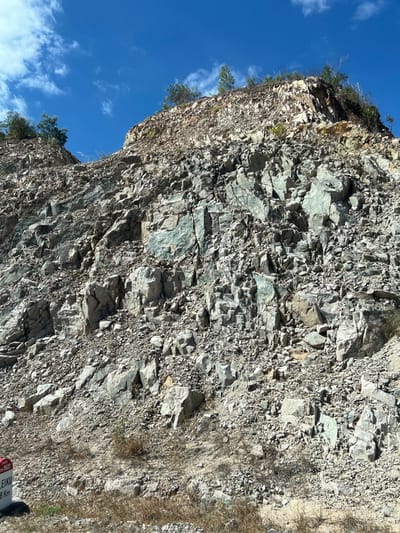 A towering rock face dominates the frame, exposing layered formations of grey and green-tinged stone. Fragments appear broken and uneven, with small plants clinging to crevices along the top edge. The sky appears in the upper left, a deep blue dotted with wispy white clouds. Dry grass and a partially visible sign are present on the right side. The perspective is angled upward, emphasizing the height and imposing scale of the cliff.