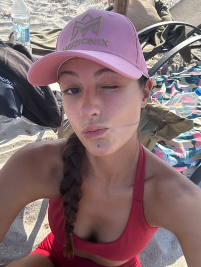 A young woman is lying on the sand, possibly at a beach, as indicated by the sand and a colorful blanket with a fruit pattern in the background. She is wearing a pink baseball cap and a red swimsuit top. Her hair is braided and resting on her shoulder. She is looking up at the camera, with one eye closed in a wink. A plastic water bottle is visible near her. The sunlight suggests it is daytime. The overall mood seems relaxed and playful. The text "MOTIONIX" is visible on the front of her cap.