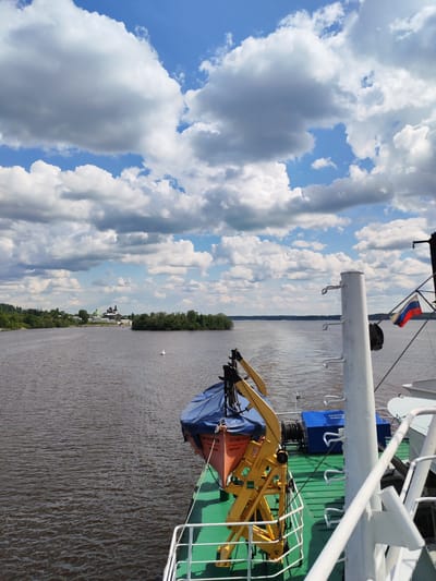 The image shows a view from a boat on a body of water, with a landmass visible in the distance under a partly cloudy sky. The boat features a lifeboat covered in blue tarpaulin, secured by a yellow support structure. A white railing and a pole with the Russian flag are present on the right side of the image. The water is a dark brown color. The buildings on the landmass in the distance are identifiable as the Kirillo-Belozersky Monastery, a significant landmark in the city of Goritsy, Russia.