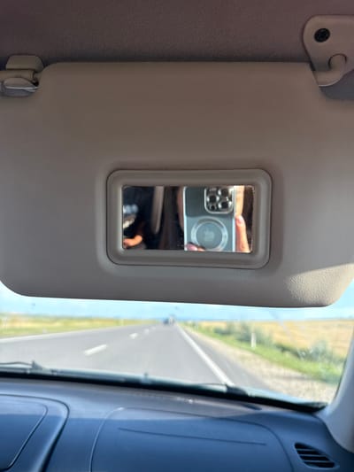 An indoor, eye-level shot taken from inside a car shows a grey sun visor with a small rectangular mirror embedded in it. A person's hand, adorned with a pink-lacquered fingernail, is reflected in the mirror, holding a silver smartphone. The smartphone's camera array and a circular Apple logo are visible in the reflection. The sun visor is open, revealing a blurred view of a road stretching into the distance. The road appears to be in a rural area, flanked by fields of yellow and green crops under a clear blue sky. The scene is likely captured in Sagaidac, Moldova.