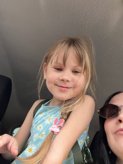 In this selfie-style photograph, two individuals, likely a mother and daughter, are captured in a car. The child, positioned in the foreground, has light blonde hair and is wearing a light blue dress adorned with a pattern of white daisies and yellow centers. A pink butterfly hair clip is visible on her hair, which is styled in a braid. Her expression is cheerful, with a slight smile. To the right and slightly behind her, the face of an adult is partially visible. They are wearing dark sunglasses and have dark hair. The background suggests the interior of a vehicle, with hints of the car's interior elements like a seat and window. The lighting appears to be natural daylight, indicating it could be daytime. The scene conveys a sense of familial warmth and a casual moment shared during a car ride, possibly within or near Малышево, Russia, given the location context. There is no visible text in the image.