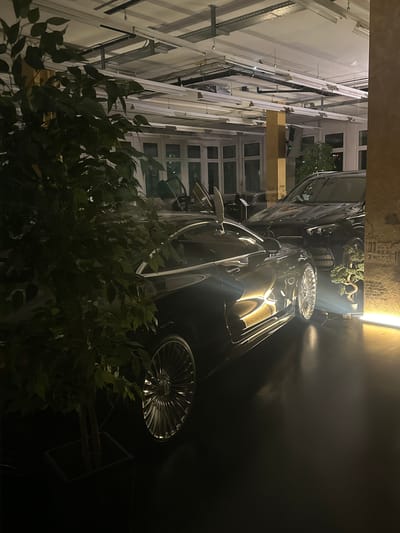 The image shows a dark-colored luxury car parked outdoors in Wetzikon (ZH), Switzerland. The car is adjacent to a building with multiple windows and what appears to be a system of exterior support structures. A plant obscures part of the car and the building. The scene is illuminated by a nearby light source, creating strong highlights on the car's surface. The time of day is likely nighttime.