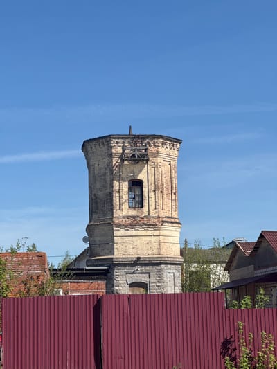The image depicts an old, partially dilapidated brick tower under a clear blue sky. It's likely the former water tower in Balezino, Russia, judging by its architectural style and the presence of similar structures in the region. The tower is mostly constructed of yellow brick with visible signs of weathering and damage, including missing bricks, particularly near the top. There's a dark window visible on one side of the tower. Wooden supports and scaffolding are present on the upper part of the tower, suggesting ongoing repairs or structural deterioration. Parts of nearby buildings are visible, including a section of a roof covered with red corrugated metal sheets and a building with a combination of brick and stone walls. Some greenery, potentially trees or bushes, is present in the foreground and around the tower.