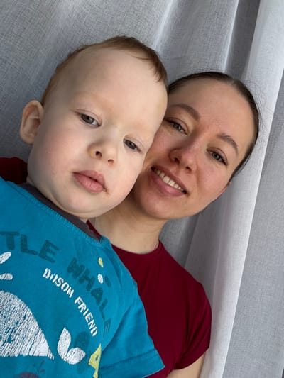 The image portrays two individuals: an adult female and a young child, positioned closely together in what appears to be an indoor setting. The adult female, on the right, has dark hair pulled back, green eyes, and is smiling with teeth visible. She wears a plain dark red short-sleeved top. The child, occupying the left half of the frame, appears to be a toddler with light hair and a neutral expression, looking directly at the camera. A small dark smudge is present above the child's upper lip. The child wears a blue short-sleeved t-shirt with a white graphic of a whale and partial text, including "LITTLE WHALE" and "OSOM FRIEND." The background is a light-colored, vertically textured fabric, consistent with a curtain or drapes. No visual elements in the image confirm the location as Gamovo, Russia.