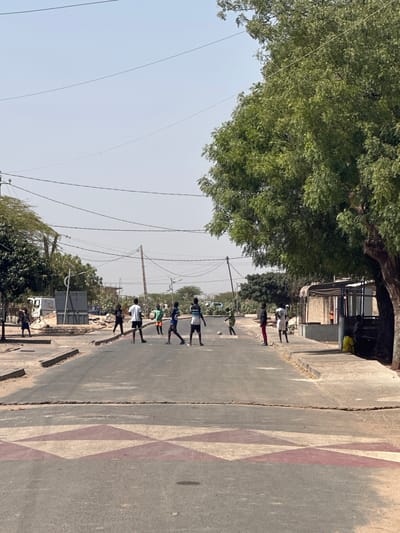 The image shows a street in the Communauté rurale de Malicounda, Senegal, with a group of people walking down the middle of the road. The road has a painted pattern on the right side. There is a large tree on the left side of the road. Buildings are visible in the background, along with telephone poles and wires. The people are spaced out along the road.