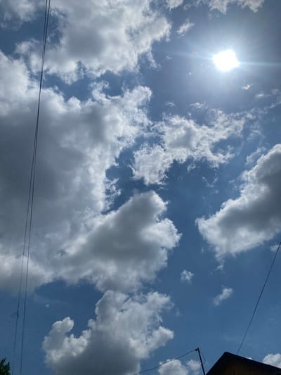 The image captures a bright, sunny day with a clear blue sky filled with fluffy white and gray clouds. The sun is visible in the upper right portion of the frame, emitting a strong glare. Several thin, dark wires, likely power lines or telecommunication cables, are stretched across the sky from the left side and the bottom right. In the bottom right corner, the dark triangular roof of a building is visible, with a pole extending upwards from it. The scene appears to be an outdoor shot looking upwards, possibly taken from a residential area in Berangas Timur, Indonesia, given the presence of utility lines and building structures common in urban or suburban environments. The overall impression is one of a typical day with overhead infrastructure against a backdrop of dynamic cloud formations.