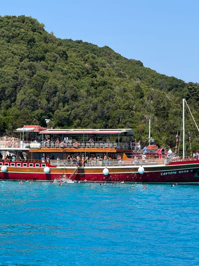 The image depicts a tourist boat named "CAPTAIN HOOK II" docked near a lush, green, tree-covered island. Several people are visible on the boat's decks, some standing, others sitting, and many are in the water swimming or playing. The boat is a multi-decked vessel with a red hull and a wooden upper structure, featuring white railings and several life preservers. The water is a vibrant turquoise blue. The background shows a steep, densely vegetated hillside under a clear blue sky. The scene suggests a recreational outing on the coast of Antipaxos, Greece, a popular destination known for its natural beauty and clear waters.