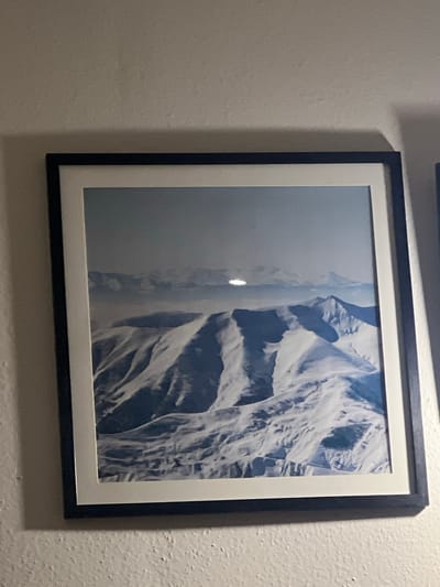 The image displays a framed photograph of snow-covered mountains. The photograph is hung on a textured, off-white wall. The frame is black with a white matting around the photograph. The photograph itself shows a vast expanse of mountains, with sharp peaks and deep valleys, all covered in snow. The sky above is a hazy blue, and there appears to be a bright light source, possibly the sun, reflecting off a distant peak. The scene depicts a remote, mountainous landscape, likely taken from an aerial perspective due to the sweeping views and the absence of any discernible human presence or structures within the image. The location is identified as Kumlistsikhe, Georgia.