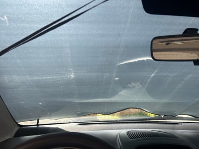 The image shows the interior of a car, looking out through the windshield. The windshield is covered by a dark grey sunshade. Two windshield wipers are visible, resting on top of the sunshade. A rearview mirror is mounted on the windshield, reflecting a brick wall and a bright light source. The dashboard is visible in the lower part of the image, with a textured steering wheel on the left and air vents on the right. Outside the car, the weather appears to be sunny, with green foliage visible beyond the windshield. The scene is likely set in Lisse, Netherlands, during the daytime.