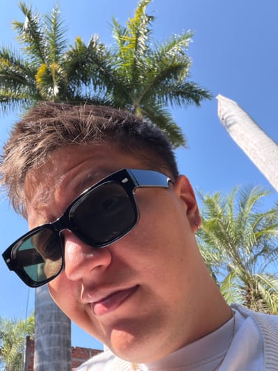 A person wearing black sunglasses looks up towards the camera. The sky and palm trees are visible in the background. The person's face is angled, with a slight smile. They are wearing a white sweater. The sunglasses reflect the surrounding environment. A bit of a white pole is also visible behind the palm tree.