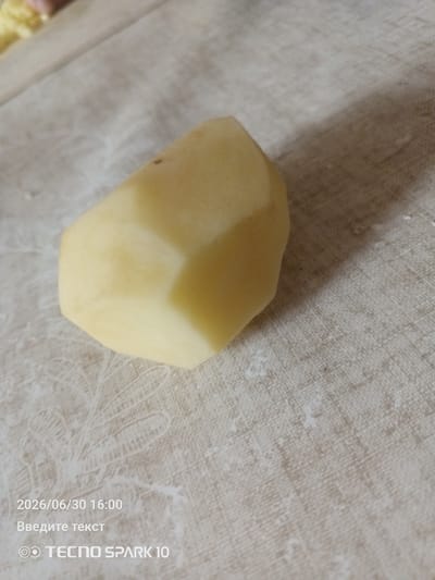 A single, peeled potato chunk is positioned on a textured, light-colored surface with a faint floral pattern. The potato is cut with angular facets, revealing its pale yellow flesh. The surface it rests on is a light beige or off-white textile, possibly a tablecloth or placemat. In the bottom left corner, text overlays the image, indicating a date and time: "2026/06/30 16:00", followed by "Введите текст" which translates to "Enter text", and "TECNO SPARK 10" with a logo. The lighting suggests an indoor setting, casting a soft shadow from the potato onto the surface. There are no people or animals visible in the image. The specific location in the city of Жанай, Kazakhstan, is not discernible from the visual information provided.