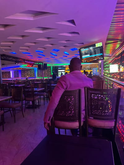 This image depicts the interior of a vibrant entertainment venue, likely a bar or lounge, located in Kimorori, Kenya. The scene suggests it is nighttime or evening, given the reliance on artificial lighting.
A man, viewed from the back, is the primary human subject. He is seated at a dark table, wearing a light-colored blazer or suit jacket. He faces a prominent white wall on the left, which features intricate geometric cut-out patterns. These patterns are backlit by striking blue lights, creating a modern and dynamic visual effect.
Numerous ornate chairs are visible throughout the venue. These chairs have distinctive patterned backs, possibly made of metal, and light-colored padded seats. Some chairs are stacked, indicating either preparation or closing hours. Multiple tables are spread across the space.
Several flat-screen televisions are mounted on the patterned wall. One screen clearly displays a list or schedule, with visible text headers including "TIME," "ROOM," "NAME," "QTY," and "DATE." Specific entries like "01:00 2023 07.24" are legible, suggesting a timestamp or event detail, with a smaller image or video playing below the text.
The venue is illuminated by a variety of colorful lights, including blue, green, and red stage lights, as well as string lights on the ceiling, contributing to a lively and energetic atmosphere. The floor is tiled with a light-colored material. The man appears to be observing his surroundings or the content on the screens, though no specific activity is clearly depicted.