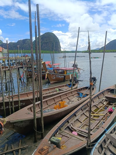 In Ko Panyi, Thailand, a waterfront scene depicts several wooden boats moored at a stilted pier. The boats are of varying sizes and appear weathered, with some containing cargo such as yellow crates and a pink umbrella. The water is murky, and wooden posts, some covered in barnacles, rise from the water to support the pier structure. In the background, lush green mountains rise from the sea, and floating fish farms are visible on the water. The sky is partly cloudy with patches of blue. The image captures a typical scene of daily life and maritime activity in this unique floating village.