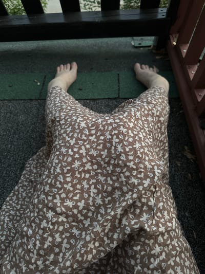 The image shows a person's bare feet and legs from a top-down perspective, wearing a brown dress with a white floral pattern. The person is sitting or lying down on a textured surface, possibly a wooden deck or patio, with some green roofing material visible beneath them. Dark wooden beams and railings are present in the background, suggesting an outdoor setting. The lighting indicates it is daytime, but potentially overcast or late in the day. The scene does not provide any specific landmarks or visual cues to determine its exact location within McMasterville, Canada.