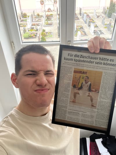 A young adult male, with short dark hair and fair skin, is depicted indoors, positioned in the foreground. He wears a light-colored, crew-neck t-shirt. His face exhibits a deliberate expression characterized by squinted eyes, a wrinkled nose, and pursed lips. He holds a black-framed document, identified as a newspaper clipping, in his right hand, presenting it to the right side of his head.

The newspaper clipping displays a prominent German headline: "Für die Zuschauer hätte es kaum spannender sein können" (For the spectators, it could hardly have been more exciting). Below the headline, the text refers to "Basketball-Bezirksliga" and "TV Memmingen." An embedded photograph within the clipping shows a male basketball player, wearing a white jersey with the number 5, light shorts, and light-colored athletic shoes. He is captured in a triumphant pose on a wooden court, with bent knees and clenched fists. A second player in a yellow jersey, partially visible, bears the number 12.

The individual is standing in front of a multi-pane window with a white frame. Through the window, an outdoor scene is visible: a cemetery. Numerous grave plots are present, each featuring a headstone or monument and diverse plantings. The ground between the plots appears to be graveled. Additional foliage is visible beyond the immediate grave plots. The sky is overcast.

In the lower right corner of the image, partially obscured by the framed clipping, folded items of clothing in black, white, and red are visible on an indoor surface, likely a shelf or dresser.

Based on the reference to "TV Memmingen" within the newspaper clipping and the provided context, the location of this scene is understood to be in Benningen, Germany, specifically Benningen (Unterallgäu), which is situated in proximity to Memmingen. The visible cemetery aligns with a community burial ground in such a German municipality.