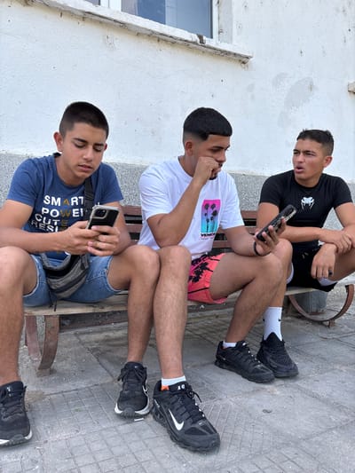 Three young men are sitting on a metal bench in Mihailovo, Bulgaria. The first man, wearing a black t-shirt and black shorts with white socks and black sneakers, sits on the upper portion of the bench, looking at his phone. The second man, wearing a white t-shirt with a palm tree design and red shorts with palm trees, touches his face, possibly looking at the phone. The third man, wearing a blue t-shirt and jean shorts with a gray fanny pack, is looking at his phone. All of them are sitting close together. The bench appears to be outside of a building with white walls. The ground is paved.