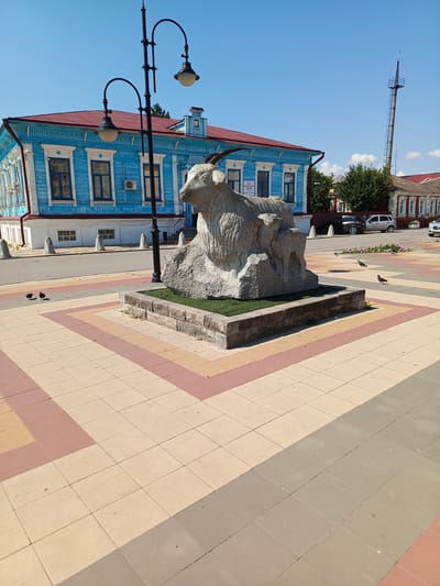 In Uryupinsk, Russia, a stone sculpture of a goat stands prominently on a paved plaza. The sculpture, crafted from light-colored stone, depicts a goat with horns and appears to be situated on a rocky base surrounded by a low stone border filled with grass. Behind the sculpture, a brightly painted blue wooden building with a red roof, featuring white trim and decorative window frames, is visible. To the right of the blue building, a smaller, older-looking building with a red roof and white window detailing is present, with cars parked in front of it. The plaza itself is paved with square tiles in a repeating pattern of beige, pink, and light brown. A double-headed lamppost stands to the left of the sculpture. Several pigeons are scattered across the paved area. In the background, a tall metal tower with a ladder extends upwards against a clear blue sky with a few wispy clouds. The overall scene suggests a public space in a town or city.