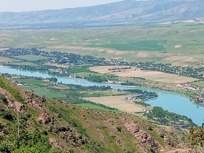 The image presents a landscape view from a high vantage point, likely a hillside, overlooking a broad valley with a river winding through it. The scene suggests the location is near or in Chinarkent, Uzbekistan, given the terrain and settlement patterns.
The foreground is dominated by a rocky hillside covered in sparse vegetation. The river is the most prominent feature in the mid-ground, exhibiting a distinct blue-green color. It meanders through the valley, bordered by agricultural fields and small settlements with dense vegetation.
In the background, rolling hills fade into the distance, culminating in a mountain range under a hazy sky. Evidence of human infrastructure is apparent in the distant landscape, including electrical transmission towers.
The settlements are small and clustered, with buildings appearing as low, dense constructions surrounded by trees. Patches of cultivated land interspersed with natural vegetation indicate an agricultural economy.