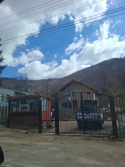 The image depicts a street scene in Gintsi, Bulgaria. A two-story house is visible behind a black metal fence. A silver car is parked in the driveway of the house. To the left of the house, a lower building with a red facade and windows is also visible behind a fence. In the background, a forested mountain slopes upward. The sky is blue with scattered white clouds. Power lines stretch across the sky in the left portion of the image. The ground is paved, appearing to be a road or sidewalk.