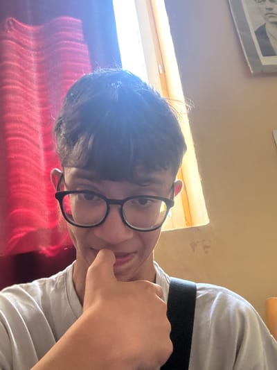 A person with dark hair and glasses is seen indoors, possibly in Obruchishte, Bulgaria, as requested. The individual is wearing a white shirt with a black band across the chest. Their hand is touching their face. A window is behind them, with a red curtain partially visible. There is a framed picture on the wall. The image is taken from a low angle.