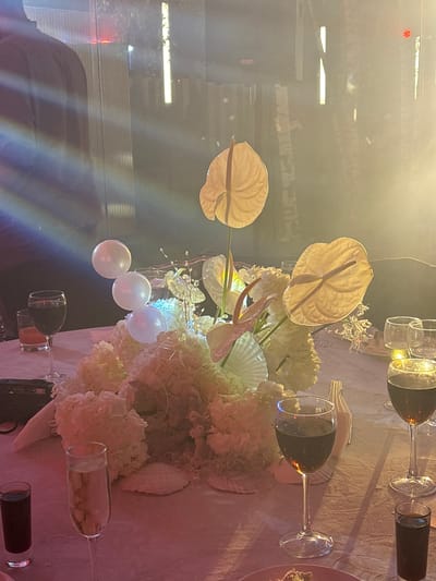 A formal event is depicted in a dimly lit room with soft, colored lighting. In the foreground, a table is set with various drinks in glasses, including red wine and champagne. A floral centerpiece dominates the center of the table, featuring large, pale anthurium flowers and a fluffy arrangement of white flowers, possibly hydrangeas. There are also decorative shells placed around the centerpiece. In the background, there are hints of people and the environment, with vertical lights and a light display creating a blurred, atmospheric effect. The overall impression is one of a festive gathering or celebration.