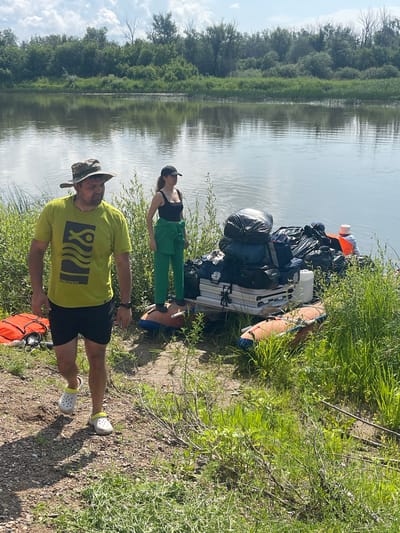 This image captures a scene by the river in the city of Nezhinka, Russia. In the foreground, a man wearing a camouflage hat, a lime green t-shirt with a graphic design, and black shorts is walking towards the viewer. He is also wearing white Crocs. To his left, on the riverbank, is an orange life vest. Behind the man, a woman stands near a large pile of gear on a small cart. She is wearing a black tank top and green pants, with a black baseball cap. The gear pile consists of several large black trash bags, duffel bags, a white plastic container, and what appear to be folded white tables. An inflatable raft lies on the ground in front of the cart. The background shows a wide, calm river reflecting a cloudy sky and a dense line of trees and vegetation along the opposite bank. The overall environment is natural and appears to be a staging area for an outdoor activity, likely involving the river.