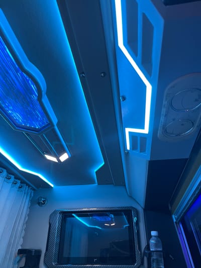 This media file depicts the interior of a vehicle, likely a luxury bus or van, with a futuristic and modern design. The dominant visual element is the pervasive blue LED lighting that outlines various features of the cabin.
The subjects are primarily the objects within the vehicle. These include what appear to be air vents or control panels on the upper part of the interior, a tablet device mounted on the wall with a carbon fiber-style frame, and a bottle of water in a holder. Below these, a section of white, pleated upholstery, possibly seats or curtains, is visible.
The scene is a static shot of the interior, with no people or activity taking place. The lighting creates a cool, ambient atmosphere. The emphasis is on the design and technology integrated into the vehicle's interior.
Notable details include the geometric patterns of the illuminated elements, particularly the sharp angles and lines of the blue lighting. The tablet screen is dark, reflecting the surrounding blue lights. The presence of the water bottle suggests a functional aspect of the vehicle, perhaps for passengers on a journey. The location cues, such as the mention of Dong Nai City, Vietnam, provide geographical context, though there are no specific visual markers within the image that definitively place it there.
There is no visible text in the image. The overall impression is one of a sophisticated and well-equipped interior, designed for comfort and a high-tech experience. The time of day and weather are not discernible from the image.
