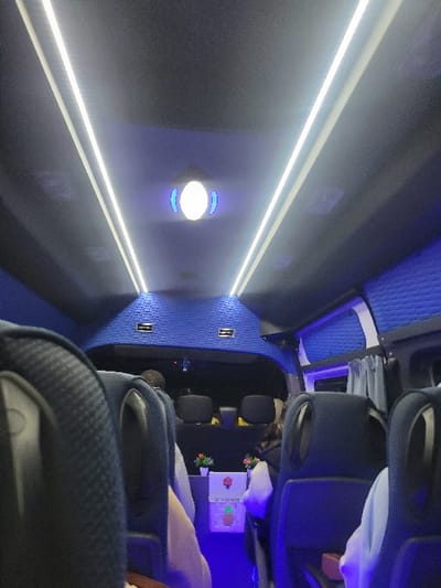 An interior shot of a passenger vehicle like a bus or limousine. Rows of dark-colored, plush seating with headrests are visible. Overhead, long, parallel light strips illuminate the space, and circular lights are mounted in the center. The ceiling and walls are blue with a textured pattern. A few passengers are seated in the rows.